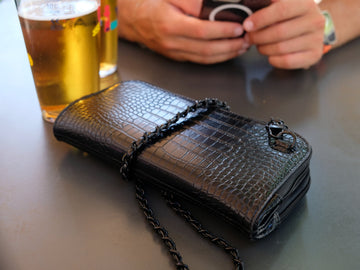 Black croc-textured Vitale by Myriad crossbody with a black chain strap on a bar table next to glasses of beer