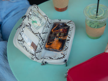 Natural snake print Vitale by Myriad crossbody open on a teal cafe table, with a phone in the large bottom pocket and cash in its own compartment
