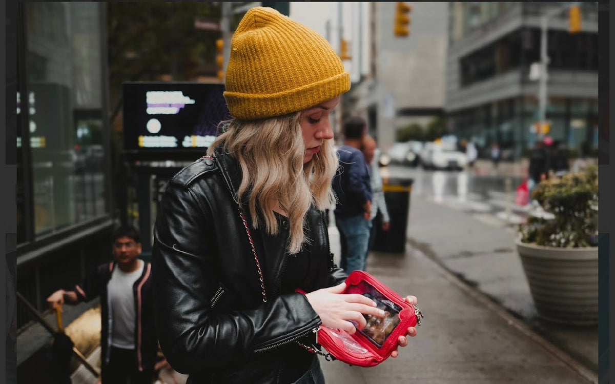 Woman in a yellow beanie using her phone through the clear window of a red Vitale by Myriad crossbody on a city street