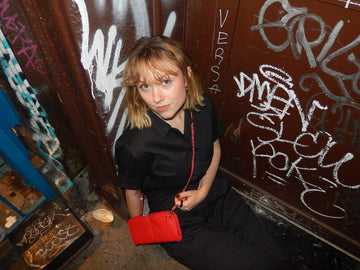 Woman in a black jumpsuit sitting against a graffiti-covered wall at night, wearing a red pebble Vitale by Myriad crossbody on a red chain strap