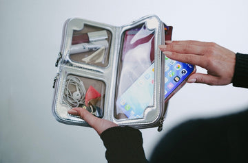 Hand sliding a phone into the large touchscreen pocket of an open silver metallic Vitale by Myriad crossbody, with earbuds and lipstick in the two smaller compartments