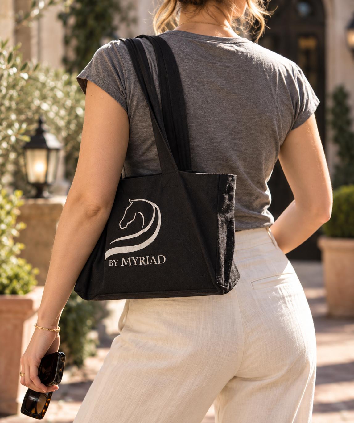 Woman carrying a black canvas tote with the by Myriad logo over her shoulder
