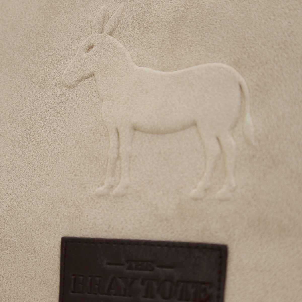 Close-up of the embossed donkey and BRAY TOTE logo tag on taupe faux suede
