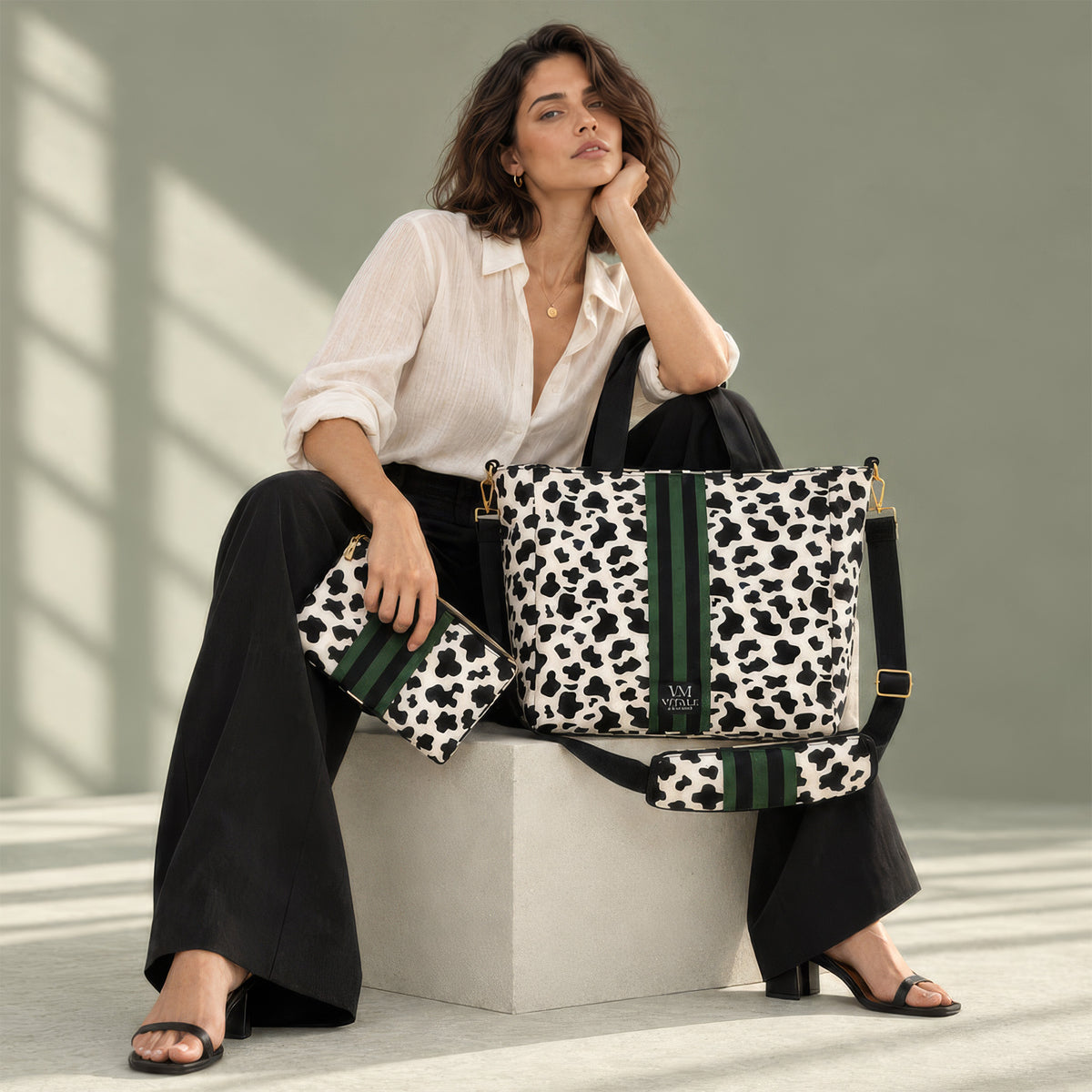 Woman in a white blouse and black trousers seated on a concrete block beside the Vitale Dublin cow print tote and its pouch