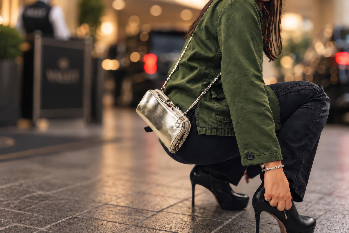 Woman in a green jacket crouching by a hotel valet stand, wearing a silver metallic Vitale by Myriad Hestia crossbody clutch