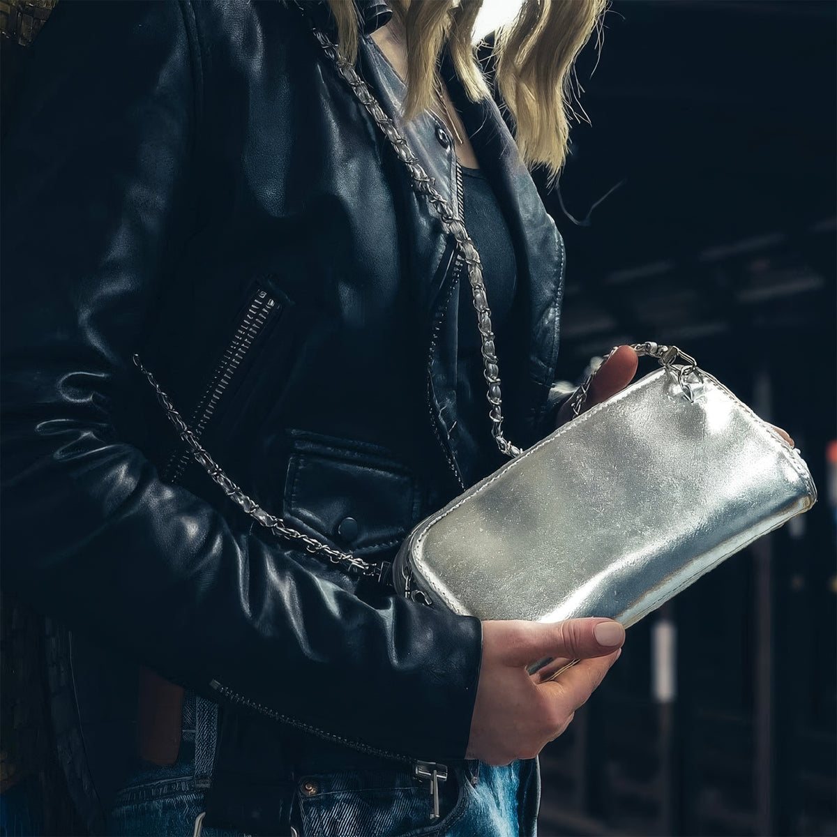 Woman in a black leather jacket holding the Hestia silver metallic mini clutch beneath an elevated train track