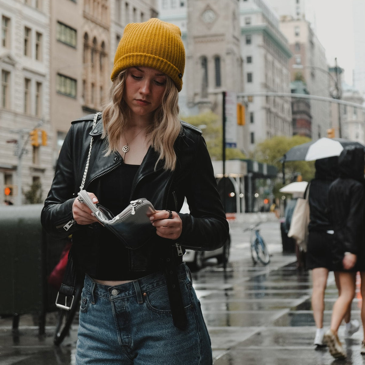 Woman in a yellow beanie wearing the Hestia silver metallic mini crossbody on a rainy New York City street
