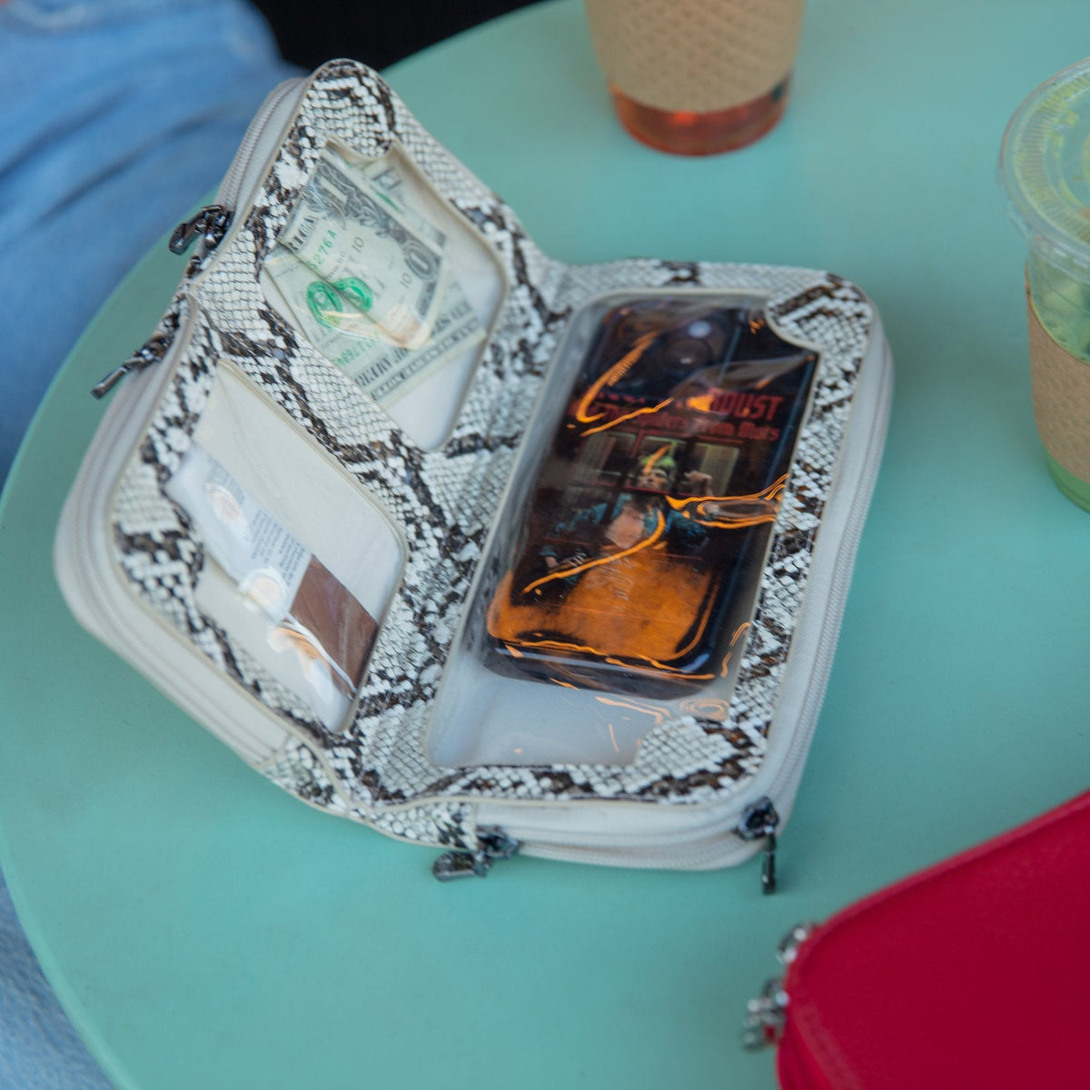 Natural snake print Vitale crossbody open on a teal cafe table, phone in the bottom pocket and cash in its own compartment