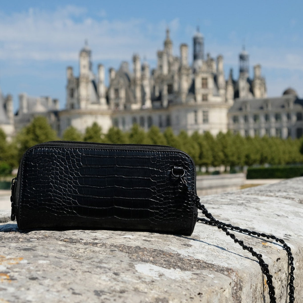 Osmand black shiny croc mini resting on a stone wall in front of a French chateau