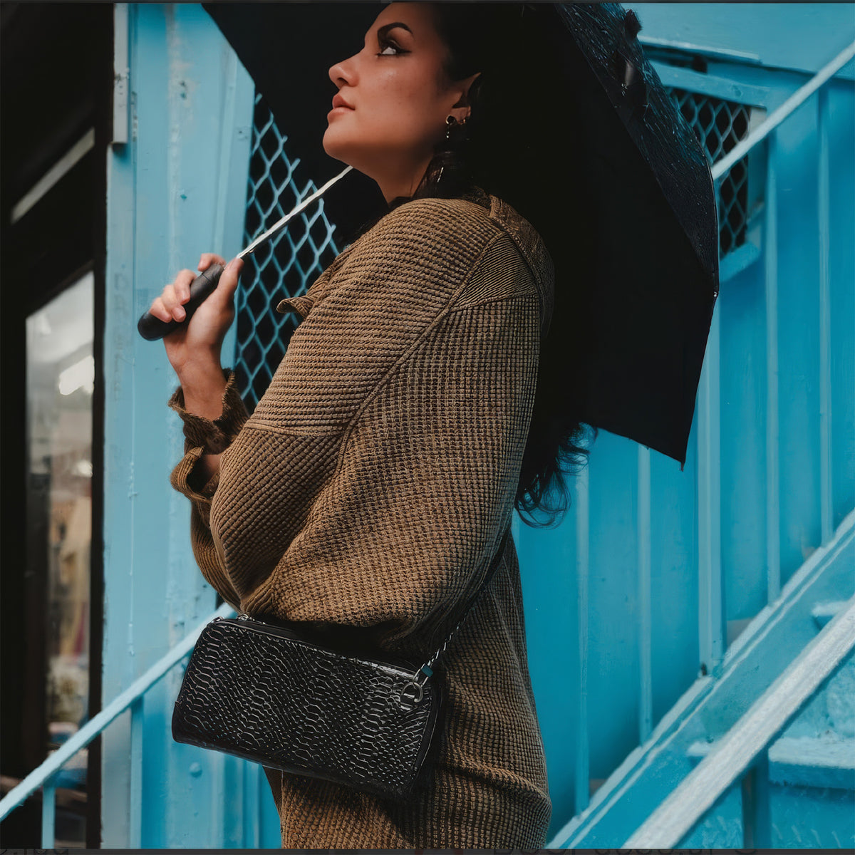 Woman with an umbrella beside blue stairs wearing the Petrel black snake mini crossbody clutch