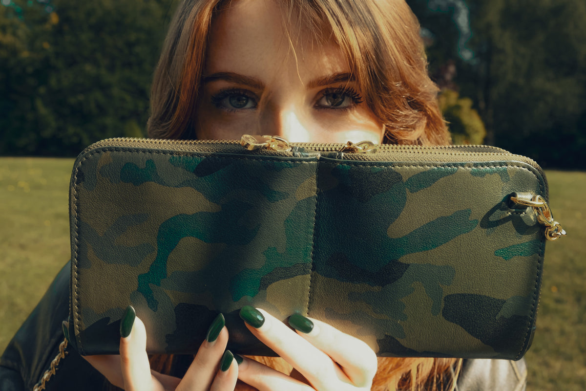 Woman holding a Zeloso green camo leather clutch with gold hardware up to her face outdoors, Vitale + House of Alimena